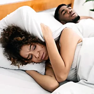 a couple in bed; the husband is snoring and the wife is covering her ears with a pillow
